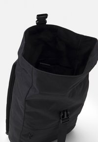 Mochila de tela negra con cierre enrollable y hebilla ajustable. El interior presenta una textura suave con un diseño minimalista y sin bolsillos visibles.