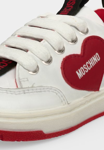 Baskets blanches avec de gros lacets blancs, semelle rouge, et un patch en forme de cœur rouge sur le côté avec "MOSCHINO" en lettres blanches.