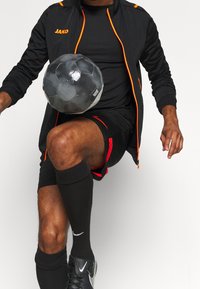 Svart idrottsjacka med oranga accenter och logotyp, svart tröja, shorts med röd trim, svarta strumpor och en svart fotboll.