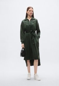 Φόρεμα χακί πράσινο με ζώνη, μακριά μανίκια, τσέπες στο στήθος με φερμουάρ και πλευρικές σχισμές. Φορεμένο με λευκά αθλητικά παπούτσια και μαύρη τσάντα.