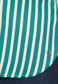 Striped skjorte med teal og hvite vertikale striper, glatt tekstur, rund hem og en liten merket logo med røde, hvite og blå detaljer.
