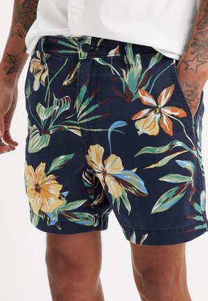 Mand iført marineblå shorts med stort tropisk blomstermønster i gul, grøn, orange og lyseblå, parret med en hvid skjorte.
