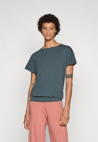 Loose-fitting, short-sleeve top in dark teal fabric with a round neck and a ribbed hem, paired with light pink wide-leg pants.