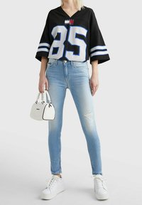 Jersey negra de gran tamaño con acentos en blanco y azul y el número "85", combinado con jeans ajustados de mezclilla azul claro con desgastes, y un bolso blanco.