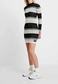 Abito a coste nero e beige con righe orizzontali, maniche lunghe, colletto alto e un patch del logo CK vicino all'orlo. Abbinato a sneakers bianche.