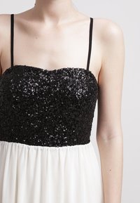 Zwarte pailletten buste met een aansluitend ontwerp, overgaand in een witte, vloeiende chiffonrok. Heeft dunne zwarte bandjes en een soepele textuur.