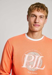 Sudadera naranja con cuello redondo blanco y mangas largas. Presenta un diseño gráfico con "PJL" y un patrón de globo en el centro.