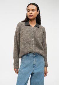 Cardigan texturat maro cu guler negru, cu închidere cu butoane și mâneci lungi, purtat cu blugi deschis la culoare, cu talie înaltă.