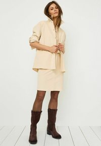 Chemise beige à boutonnières, jupe beige ajustée et bottines en daim marron foncé à talon carré, mettant en avant une texture lisse.
