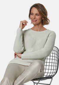 Lichtgrijze gebreide trui met ribbelstructuur, ronde halslijn en lange mouwen, gecombineerd met glanzende beige broek, zittend op een draadstoel.