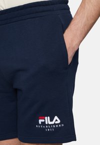 Marineblauwe katoenen shorts met zijzakken, elastische tailleband en witte geborduurde "FILA" logo met rode en blauwe accenten.