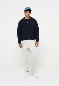 Donkerblauwe gebreide trui met een kwart-ritskraag en een klein logo, gecombineerd met lichtbeige broek en witte sneakers. Blauwe pet op het hoofd.
