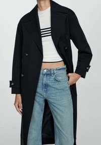 Longue veste noire en tissu lisse, avec fermeture à double boutonnage et larges revers, associée à un haut court blanc et un jean bleu clair.