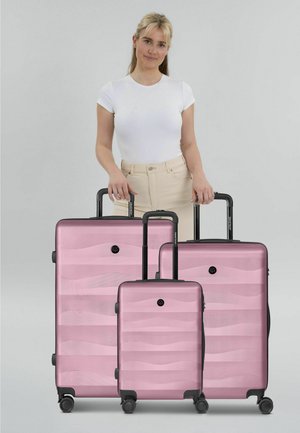 Vrouw in wit overhemd en beige broek staand achter drie roze harde koffers met zwarte handvatten en wielen op een grijze achtergrond.