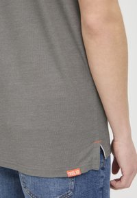 Grå t-shirt med rund halsudskæring, korte ærmer og et orange mærke i kanten. Stoffet har en blød tekstur og sideslidser.