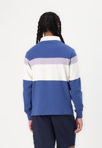 Camisa de rugby de mangas longas em azul, branco e listras lavanda. Apresenta um decote com colarinho e um corte casual com uma textura suave de algodão.