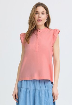 Kvinde iført en ærmeløs koralbluse med flæseærmer og et lyseblåt lagdelt skørt, stående foran en ensfarvet baggrund.