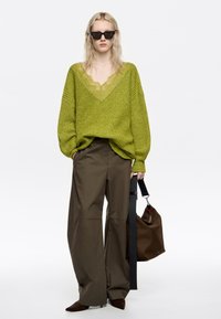 Zielony sweter dziergany z głębokim dekoltem w koronki, zestawiony z szerokimi brązowymi spodniami. Modelka trzyma brązową torbę, nosząc czarne okulary przeciwsłoneczne.