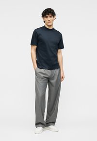 Άνδρας που φοράει σκουρόχρωμο μπλε κοντομάνικο πουκάμισο, φαρδιά γκρι παντελόνια, λευκά αθλητικά παπούτσια, στέκεται με τα χέρια στις τσέπες μπροστά σε ένα μονόχρωμο φόντο.