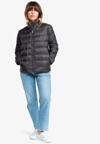 Svart puffjacka med horisontell quiltad design, ståkrage, dragkedja fram och sidofickor; bärs med ljusblå jeans och vita sneakers.
