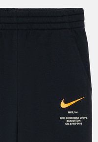 Černé bavlněné teplákové kalhoty s elastickým pasem, s oranžovým logem Nike a bílým textem potištěným na dolní části nohy.