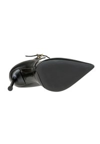 Scarpa nera con punta appuntita e tacco alto a stiletto, dotata di una sottile cinghia alla caviglia con piccola fibbia dorata, vista dal lato della suola.