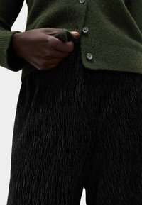 Zielony wełniany kardigan z zapięciem na guziki; teksturowane czarne spodnie z falistym wzorem; ręka umieszczona blisko kieszeni.
