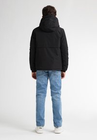 Giacca con cappuccio nera dal design elegante, abbinata a jeans blu chiaro e sneakers bianche, vista da dietro su uno sfondo semplice.