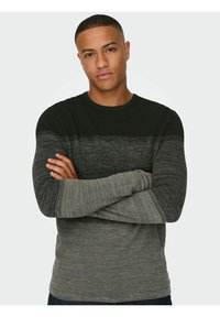 Hombre con cabello corto vistiendo un suéter de punto de manga larga con bloques de color negro, gris oscuro y gris claro en disposición horizontal, con los brazos cruzados.