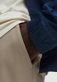 Mornarica windbreaker s elastičnim manžetama, uparen s bež majicom i hlačama. Vidljiva teksturirana tkanina. LULULEMON logo izvezen na jakni.