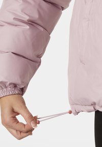 Light pink puffer jacket with elastic cuffs. A hand pulls a drawstring for adjustment, revealing soft, smooth fabric and subtle sheen.