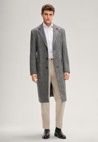 Cappotto lungo in misto lana grigio con rever a incavo, due tasche frontali e bottoni neri, indossato sopra una camicia bianca e pantaloni beige.