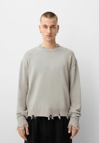 Hellgrauer gestrickter Pullover mit Rippenbund und -abschlüssen. Ausgefranste Kanten am Halsausschnitt und an den Ärmeln. Lockere Passform mit überschnittenen Schultern.