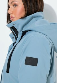 Chaqueta impermeable azul claro con cierre frontal de cremallera, capucha grande y detalles en negro. Entre los detalles destacables, un parche con la marca en la manga.