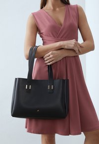 Black leather tote bag with long handles, featuring gold-tone accents and a smooth texture. Worn by a person in a pink dress.