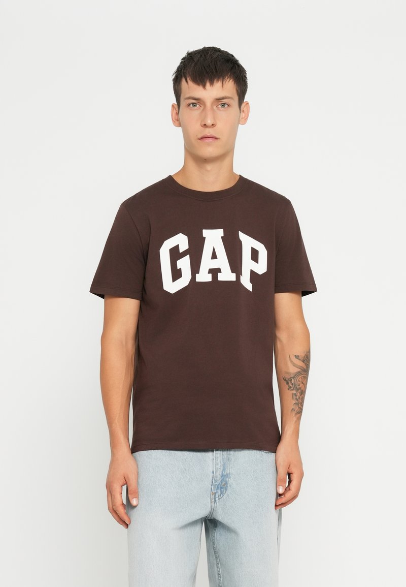 Camiseta de algodón marrón con grandes letras blancas "GAP", escote redondo y mangas cortas, combinada con jeans de mezclilla de color azul claro.