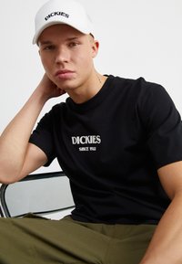 Camiseta de algodón negra con "DICKIES SINCE 1922" impreso en blanco. Gorra blanca con logotipo bordado. El modelo posa con la mano en el cuello, sentado.