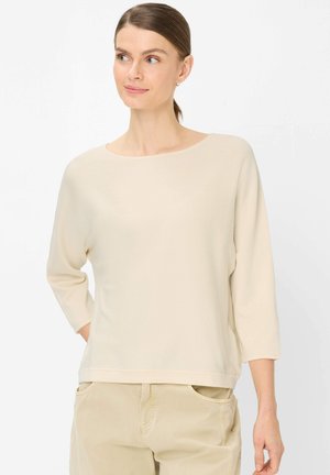 Kvinde med trukket brunt hår, iført en cremefarvet top med 3/4 ærmer og beige bukser, ser let mod sit venstre med et blidt smil.