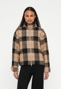 Kariertes Jackett in Beige und Schwarz mit hohem Kragen, Frontreißverschluss und zwei Vorderaschen. Hergestellt aus strukturiertem Stoff.