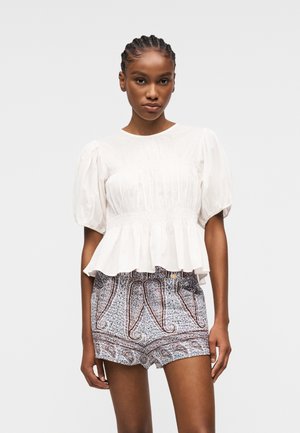 Frau trägt eine weiße Bluse mit Puffärmeln und tailliertem Schnitt sowie gemusterte, hochgeschnittene Shorts, steht vor einem schlichten Hintergrund.