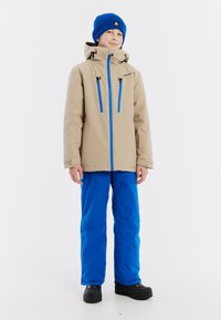 Beige geïsoleerde jas met blauwe accenten, ritszakken en capuchon; gecombineerd met blauwe skibroek en zwarte waterdichte laarzen.