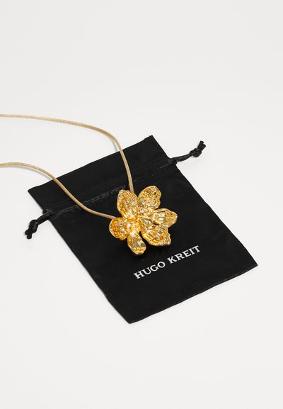 Pendente a forma di fiore in oro su una corda beige, su uno sfondo di un sacchetto a coulisse nero con la scritta "HUGO KREIT" stampata in bianco.