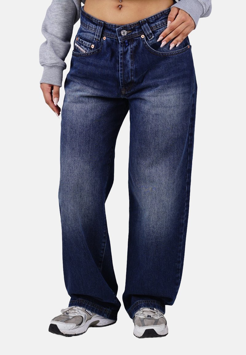 Denim wide-leg jeans in dark blue with subtle fading. Features copper hardware, front pockets, and a relaxed fit. Paired with gray sneakers.