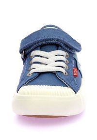 Zapatilla de lona azul con cordones blancos, tira de Velcro y suela de goma blanca. Presenta un upper texturizado y un detalle del logo rojo en el lateral.