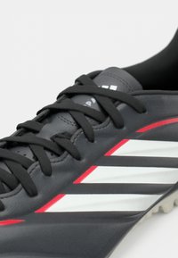 Μαύρα συνθετικά παπούτσια ποδοσφαίρου με κόκκινες και λευκές διαγώνιες ρίγες, διαθέτουν μαύρες κορδόνες και υφασμάτινη υφή στην επάνω πλευρά.