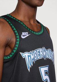 Camisola de basquetebol preta com um padrão de houndstooth verde na gola, apresentando o logotipo "TIMBERWOLVES" em azul e branco, tamanho "5."