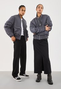 Grijze bomberjacks met geribbelde manchetten, met ritszakken; model in zwarte jeans met een middellange rok en zwarte laarzen.
