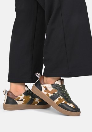 Sneakers con una combinazione di stampa di mucca e pelle nera, lacci verdi e un accento in pelle marrone sul tallone. Suola in gomma, design casual e sportivo.