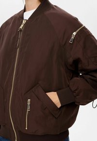Hnědá bomber bunda z hladké tkaniny, s pozlacenými zipy, žebrovanými manžetami a zipovou kapsou na rukávu. Uvolněný střih s bočními kapsami.
