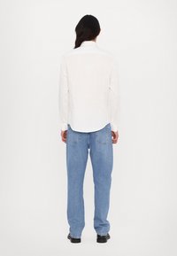 White long-sleeve shirt with a pointed collar, featuring a straight back and curved hem, paired with light blue loose-fit jeans.